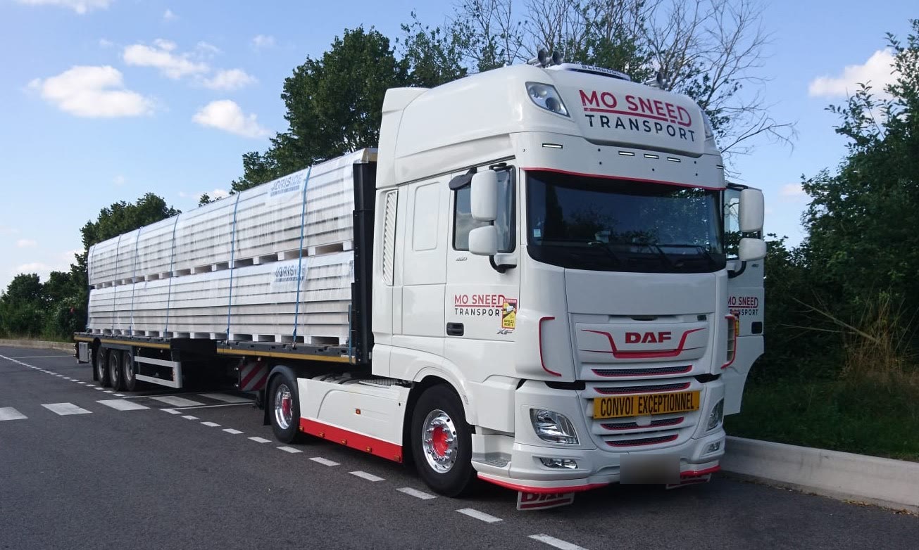 Een trekker van Mo Sneed Transport met een vlakke oplegger en een bord Convoi exceptionnel